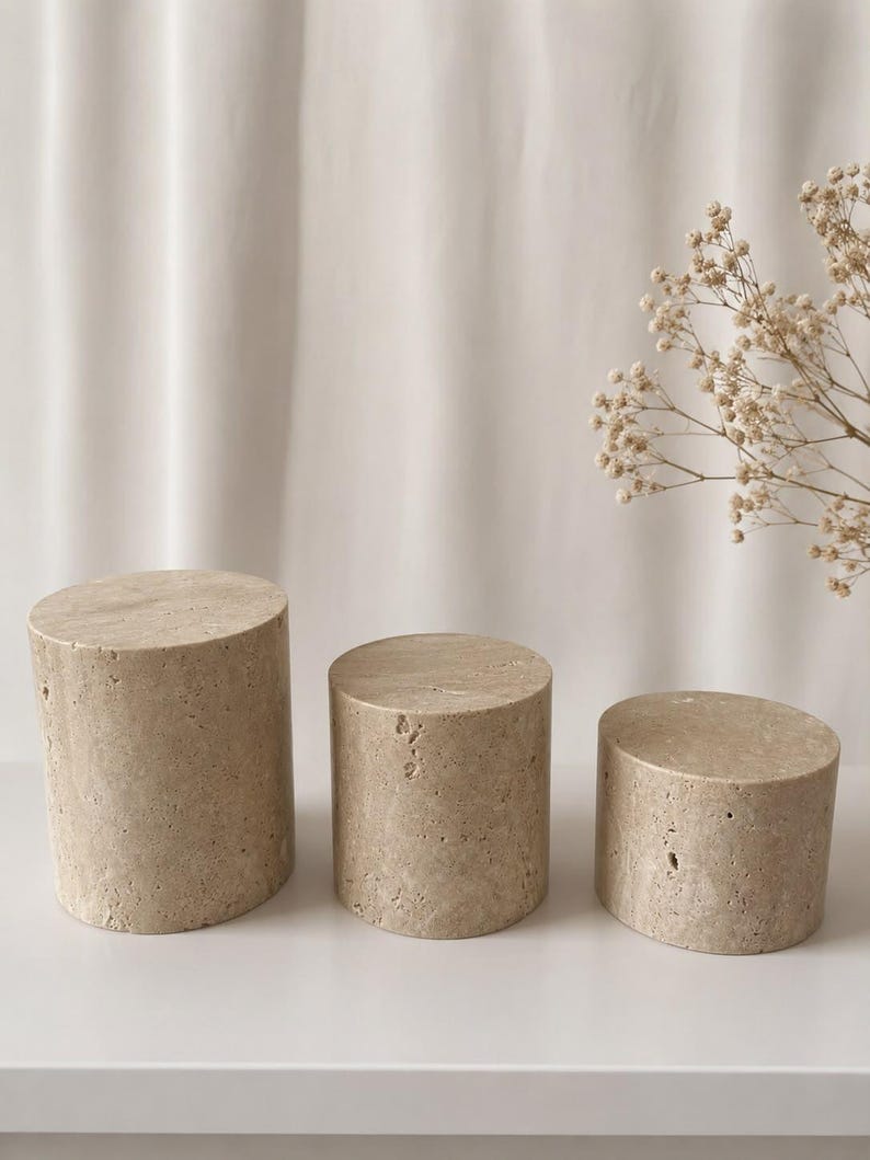 Puede incluir: Tres pedestales de exhibici&oacute;n cil&iacute;ndricos hechos de piedra travertino beige con una textura natural porosa. El conjunto incluye tres piezas de diferentes alturas, cada una con una superficie superior circular plana y una construcci&oacute;n de piedra s&oacute;lida.