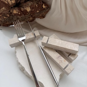 Puede incluir: Un juego de tenedor y cuchillo de acero inoxidable sobre una superficie de piedra blanca. Los utensilios tienen mangos largos y elegantes. Varios bloques de piedra rectangulares pequeños están dispuestos alrededor de la cubertería. El fondo incluye una roca grande y texturizada y tela drapeada.