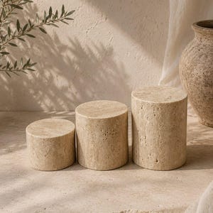 Puede incluir: Tres pedestales de exhibici&oacute;n cil&iacute;ndricos hechos de piedra travertino beige con una textura porosa natural. El conjunto incluye tres piezas de diferentes alturas, dispuestas en fila, con un color tostado claro y una superficie org&aacute;nica con poros.