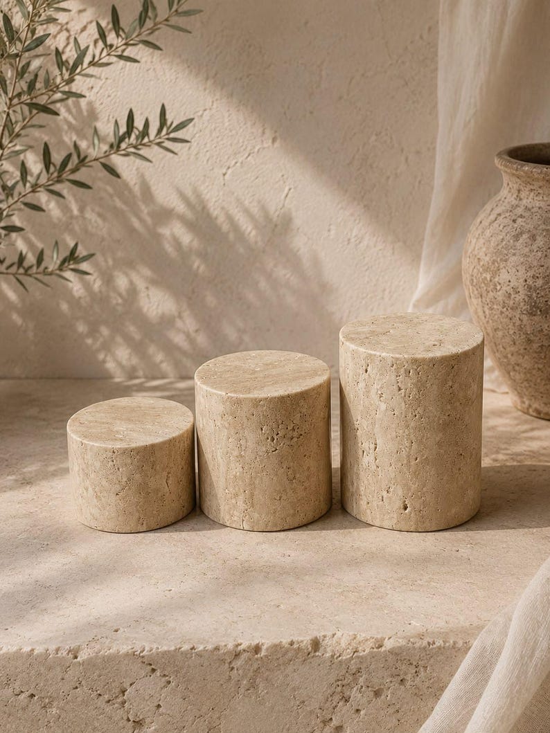 Puede incluir: Tres pedestales de exhibici&oacute;n cil&iacute;ndricos hechos de piedra travertino beige con una textura porosa natural. El conjunto incluye tres piezas de diferentes alturas, dispuestas en fila, con un color tostado claro y una superficie org&aacute;nica con poros.