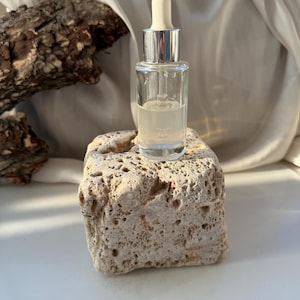 Puede incluir: Un frasco cuentagotas de vidrio transparente con tapón plateado y cuentagotas blanco, lleno de un líquido claro. El frasco está sobre una piedra beige texturizada. El fondo incluye una tela blanca y un trozo de madera oscura.