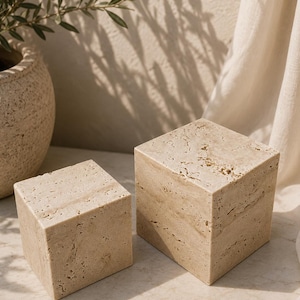 May include: Two beige travertine stone cubes with a porous, natural texture. The cubes are solid, geometric shapes with visible surface variations, pits, and veining characteristic of natural stone, displayed in two different sizes.
