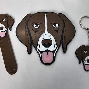 May include: A set of three brown and white dog head-shaped keychains and bookmarks. The dog is a German Shorthaired Pointer with a white patch on its chest and a pink tongue sticking out.