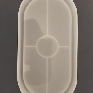 May include: White silicone mold for resin casting. The mold is oval shaped with a circular indentation in the center and four smaller rectangular indentations around the center circle.