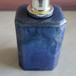 May include: A square, dark blue perfume bottle with a silver spray top. The bottle has a glittery, textured finish. The top is a shiny silver color. The bottle is a unique shape and design.