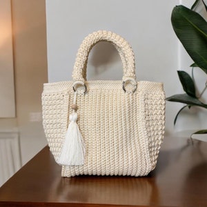 May include: A cream-colored crocheted tote bag with a white tassel. The bag has two silver rings on the top and a wooden bead on the tassel.