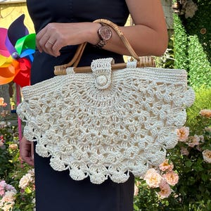 May include: Cream-colored crochet handbag with a fan-shaped design. The bag features a wooden handle and a button closure. The bag is held by a person wearing a black dress, with a watch on their wrist.
