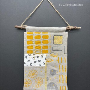 May include: A wall hanging made of linen fabric with a patchwork design. The fabric is a light beige color with yellow and gray embroidery. The hanging is attached to a branch with twine.