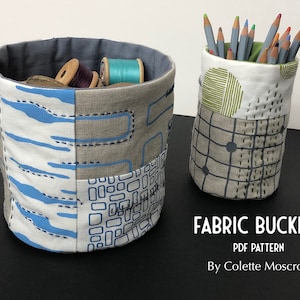 May include: Two fabric buckets, one with a blue and gray geometric pattern and the other with a green and gray geometric pattern. The buckets are filled with yarn and colored pencils. The text "FABRIC BUCKETS PDF PATTERN By Colette Moscrop" is printed below the buckets.