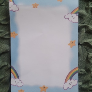 May include: A sheet of stationery with a light blue border featuring cartoon rainbows, smiling clouds, and yellow stars. The stationery has a white centre for writing or drawing.