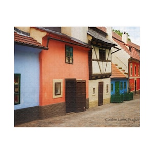 Puede incluir: Una calle estrecha bordeada de casas coloridas en Praga, República Checa. Las casas están pintadas en tonos de azul, naranja y amarillo. La calle está adoquinada y hay plantas verdes que crecen a los lados de la calle. El texto "Golden Lane, Prague" es visible en la parte inferior de la imagen.