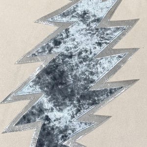 Puede incluir: Aplicación de rayo de terciopelo gris con borde metálico plateado. La aplicación está centrada en un fondo de tela beige. El rayo tiene un aspecto texturizado y está hecho de un material suave y afelpado.