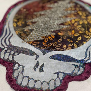 May include: A decorative skull-shaped item with a silver face and a dark purple velvet border. The center features a lightning bolt design made of silver glitter, surrounded by a colorful pattern of orange and yellow leaves.