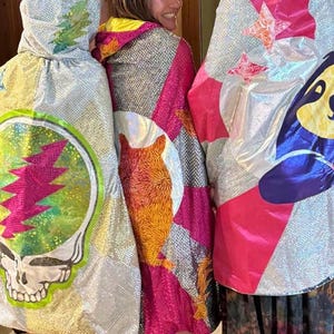May include: Three sequined capes with colorful designs. One features a skull with a lightning bolt, another has an orange bear, and the third has a cartoon character. The capes have hoods and are made of shiny fabric.