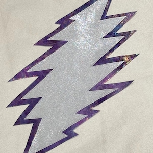 Puede incluir: Un rayo plateado brillante con un borde iridiscente púrpura y dorado. El rayo es un símbolo clásico, a menudo utilizado en la moda y el diseño. La imagen está sobre un fondo neutro.