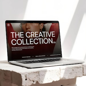 May include: A silver laptop displaying a website with the text "THE CREATIVE COLLECTION CO." on a white brick surface. The website's background is a dark red with a woman's face. The laptop is open.