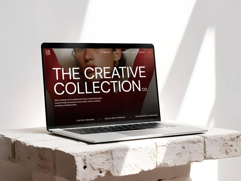 May include: A silver laptop displaying a website with the text "THE CREATIVE COLLECTION CO." on a white brick surface. The website's background is a dark red with a woman's face. The laptop is open.