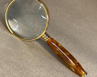 May include: A gold-toned magnifying glass with a clear lens and a marbled brown and gold handle. The magnifying glass has a decorative gold ring around the lens and at the base of the handle.