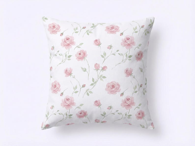 May include: Square white cushion featuring a repeating pattern of pink roses and green stems. The watercolour-style floral design includes full blooms and buds, creating a soft, romantic aesthetic. The cushion is a decorative home accent.