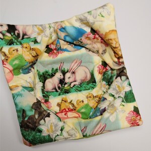 May include: A square, quilted bowl cosy with a colourful Easter-themed print. The fabric features images of bunnies, chicks, flowers, and Easter eggs in a pastel colour palette. The cosy is designed to hold a bowl of hot food.