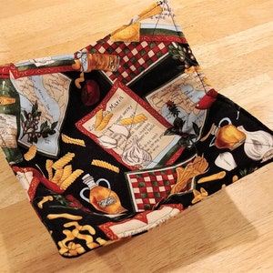 May include: A black fabric bowl with a red, white, and green Italian-themed print. The print features pasta, garlic, olive oil bottles, and a map of Italy. The bowl is folded and sitting on a wooden surface.