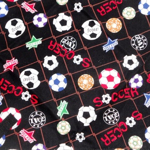 May include: Black fabric with a soccer ball pattern. The design includes soccer balls in various colors, the word "SOCCER" in red, and star-shaped graphics with "FREE KICK" and "SCORE!" text. A black strap is visible.