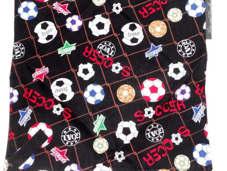 May include: Black fabric with a soccer ball pattern. The design includes soccer balls in various colors, the word "SOCCER" in red, and star-shaped graphics with "FREE KICK" and "SCORE!" text. A black strap is visible.