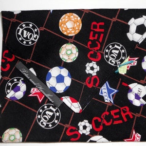 May include: Black fabric item with a soccer ball pattern, featuring soccer balls in various colors and the word "SOCCER" in red. Includes the text "GOAL" and "FREE KICK". A small label reads "Totes and Handbags".