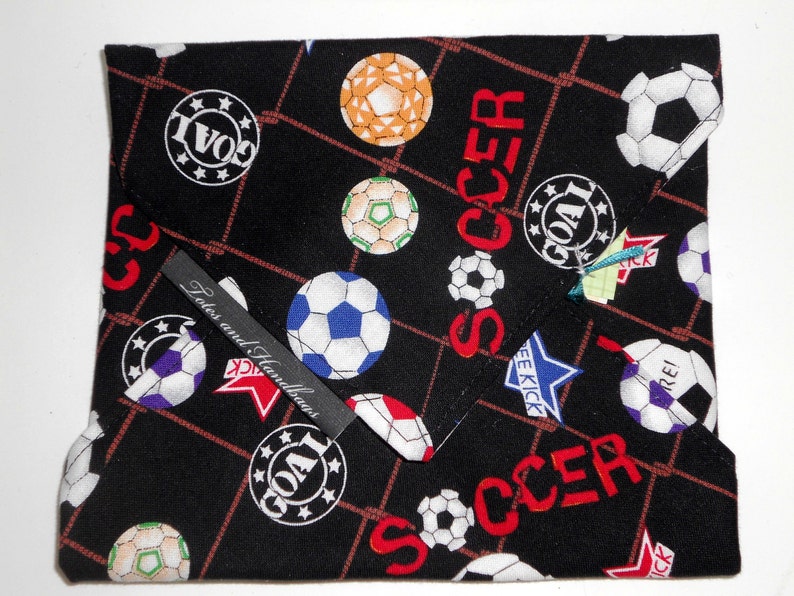 May include: Black fabric item with a soccer ball pattern, featuring soccer balls in various colors and the word "SOCCER" in red. Includes the text "GOAL" and "FREE KICK". A small label reads "Totes and Handbags".