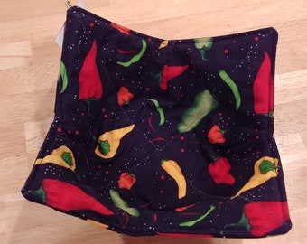 May include: A navy blue bowl cozy featuring a vibrant pattern of red, yellow, and green chili peppers. The peppers are scattered across a dark background with small white dots. The cozy is square-shaped and quilted.