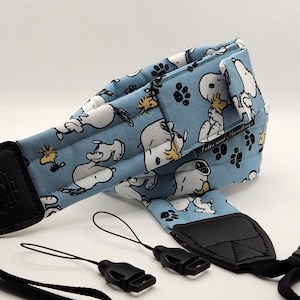 May include: A light blue camera strap featuring a repeating pattern of Snoopy and Woodstock characters. The strap has black leather ends and comes with black connector straps. The text "Lotes and Handbags" is visible on the strap.