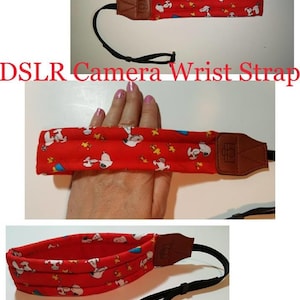 May include: Red camera wrist strap with a black adjustable strap and a brown leather pad. The strap features a repeating pattern of cartoon dogs wearing hats.