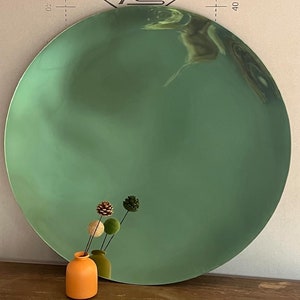 May include: A large, round, green mirror with a slightly rippled surface. A small orange vase with three small, brown and green pine cone-shaped decorations is in front of the mirror.