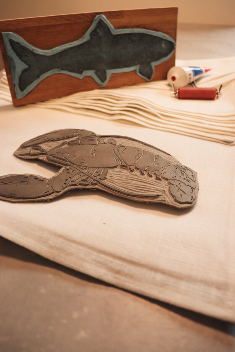 May include: A close-up of a wooden carving of a lobster. The carving is detailed and shows the lobster's claws, body, and tail. The carving is on a white surface.