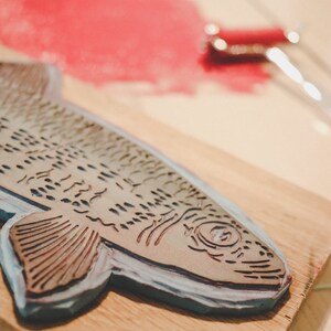 May include: A wooden block with a carved fish design, used for printing. The fish is detailed with scales and fins, and the block is painted a light blue color.