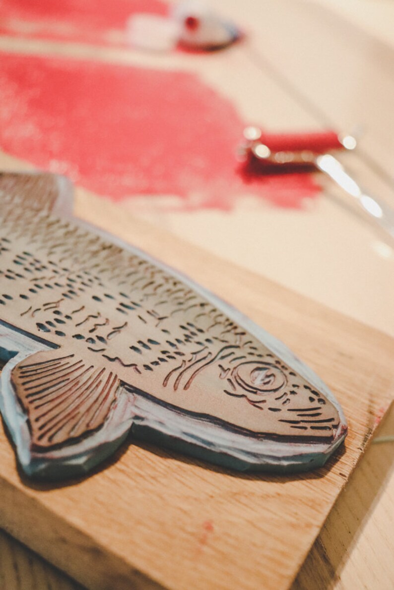 May include: A wooden block with a carved fish design, used for printing. The fish is detailed with scales and fins, and the block is painted a light blue color.