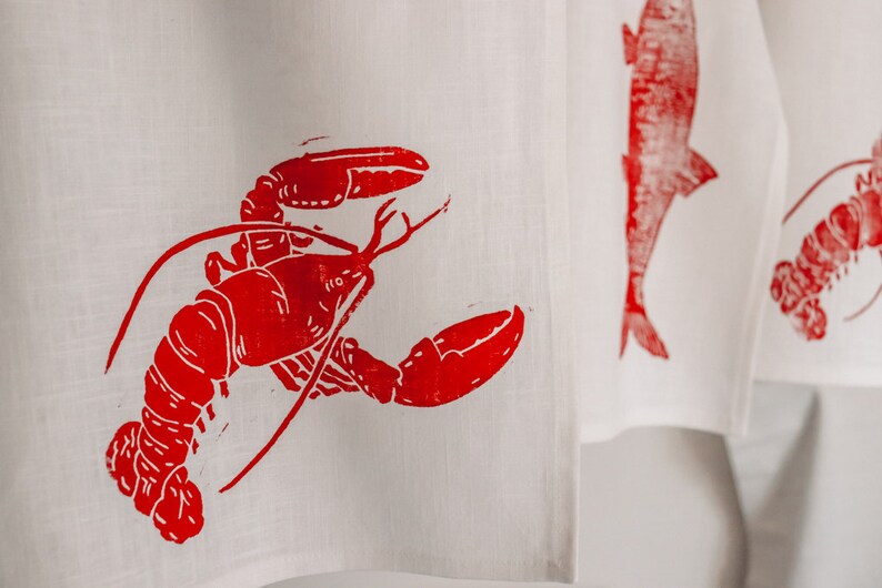 May include: A white kitchen towel with a red lobster print. The lobster is in a curled position with its claws raised.