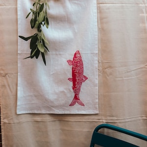 May include: A white tea towel with a pink fish design hanging on a clothesline. The tea towel is hanging on a beige background.