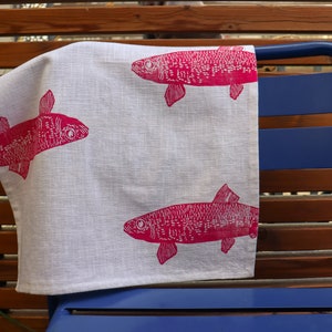 May include: A white linen tea towel with a pink fish design. The towel is hanging on a blue metal chair.