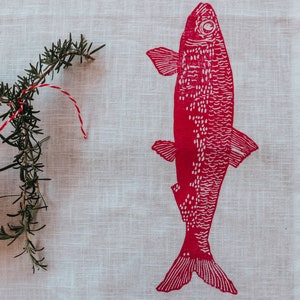 May include: A white linen kitchen towel with a red fish print. The fish has a white outline and is facing to the right. A sprig of rosemary is on the left side of the towel.