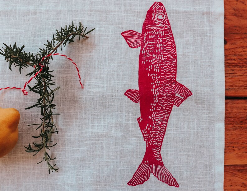 May include: A white linen kitchen towel with a red fish print. The fish has a white outline and is facing to the right. A sprig of rosemary is on the left side of the towel.