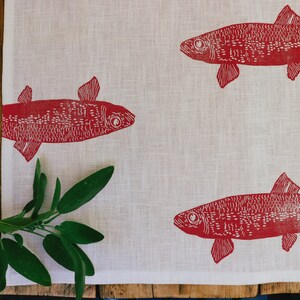 May include: A white linen tea towel with a red fish design printed on it. The fish are stylised and have a detailed pattern.