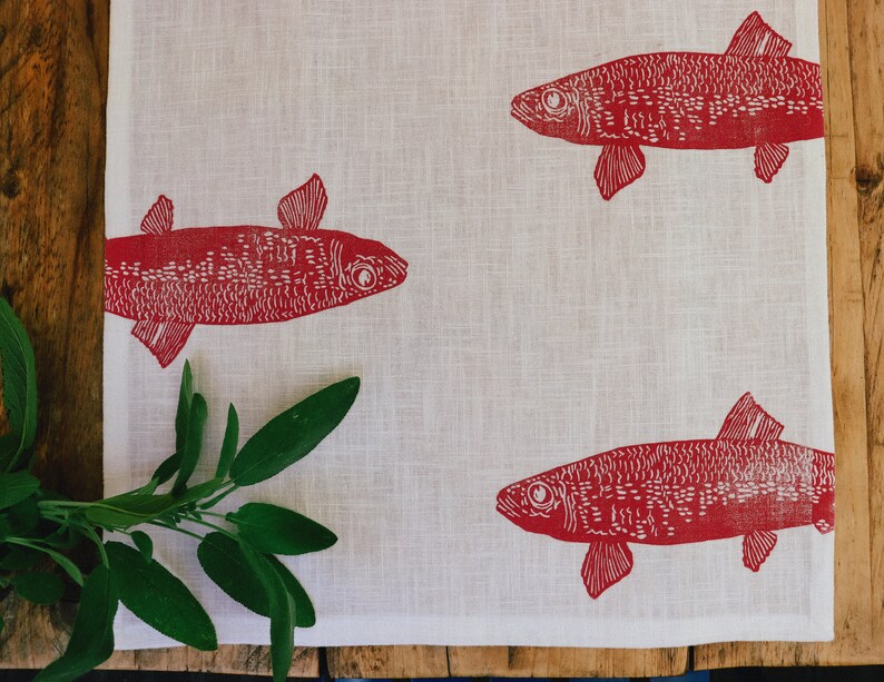 May include: A white linen tea towel with a red fish design printed on it. The fish are stylised and have a detailed pattern.