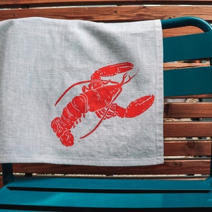 May include: A white linen tea towel with a red lobster print. The towel is hanging on a blue metal chair.
