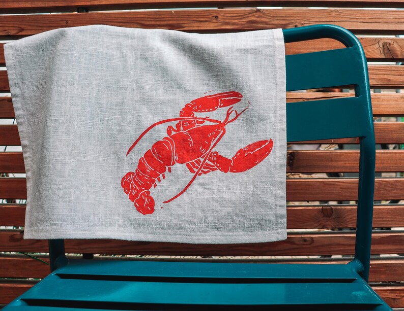May include: A white linen tea towel with a red lobster print. The towel is hanging on a blue metal chair.