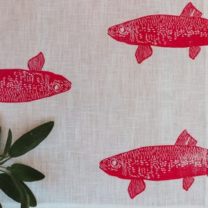 May include: A white linen tea towel with a red fish design printed on it. The fish are stylised and have a dotted pattern. A sprig of green sage is in the bottom left corner.