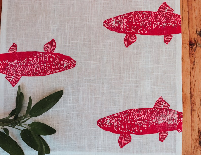 May include: A white linen tea towel with a red fish design printed on it. The fish are stylised and have a dotted pattern. A sprig of green sage is in the bottom left corner.