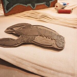May include: A close-up of a wooden carving of a lobster, with detailed lines and textures. The carving is on a white fabric surface.