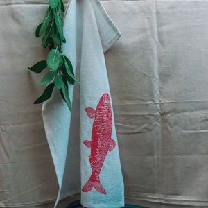May include: A white kitchen towel with a pink fish design hanging on a clothesline. A sprig of green sage is hanging next to the towel.