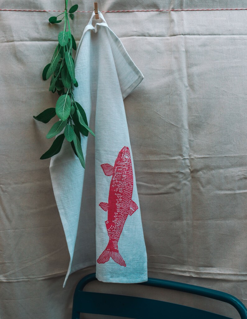 May include: A white kitchen towel with a pink fish design hanging on a clothesline. A sprig of green sage is hanging next to the towel.